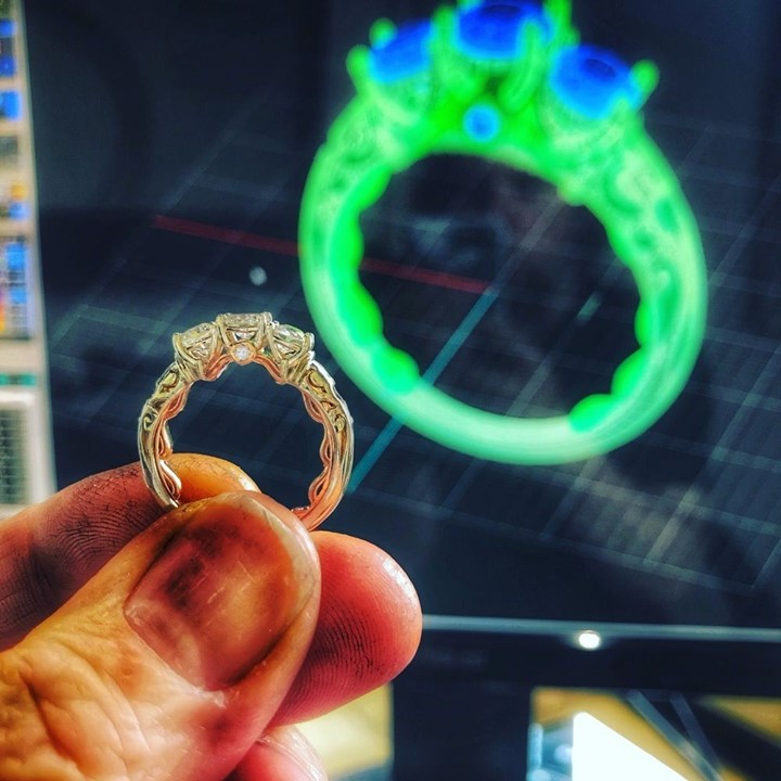 A hand holds a silver-toned ring with three clear stones in front of a computer screen displaying a green 3D model of the ring design.